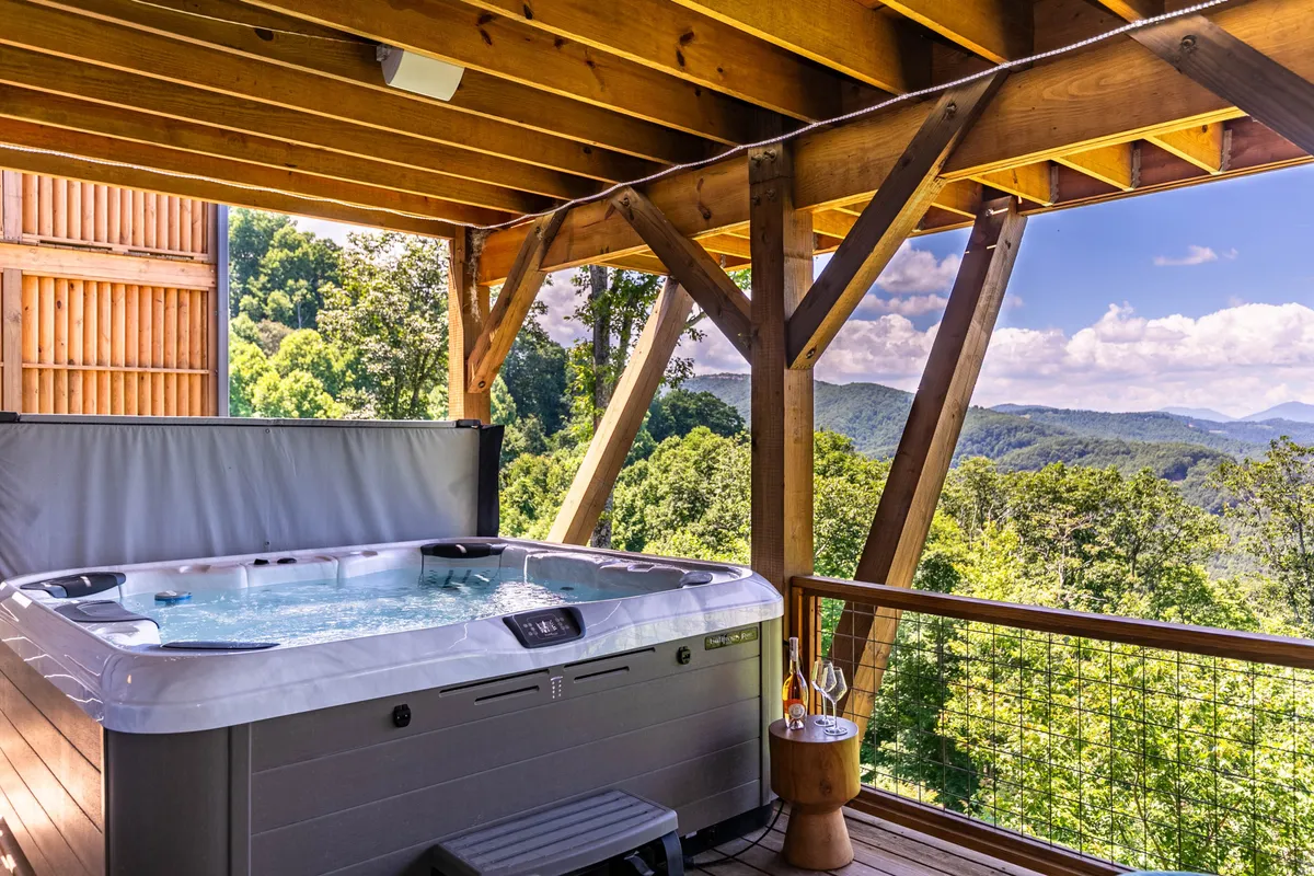 An outdoor hot tub sits under sturdy wooden beams, offering a panoramic mountain view. The late-afternoon sunlight hints at the possibility of both sunset soaks and stargazing. There are touches of luxury and serenity in the setting, with room for couples or a small group to relax in the bubbling water while surrounded by nature.