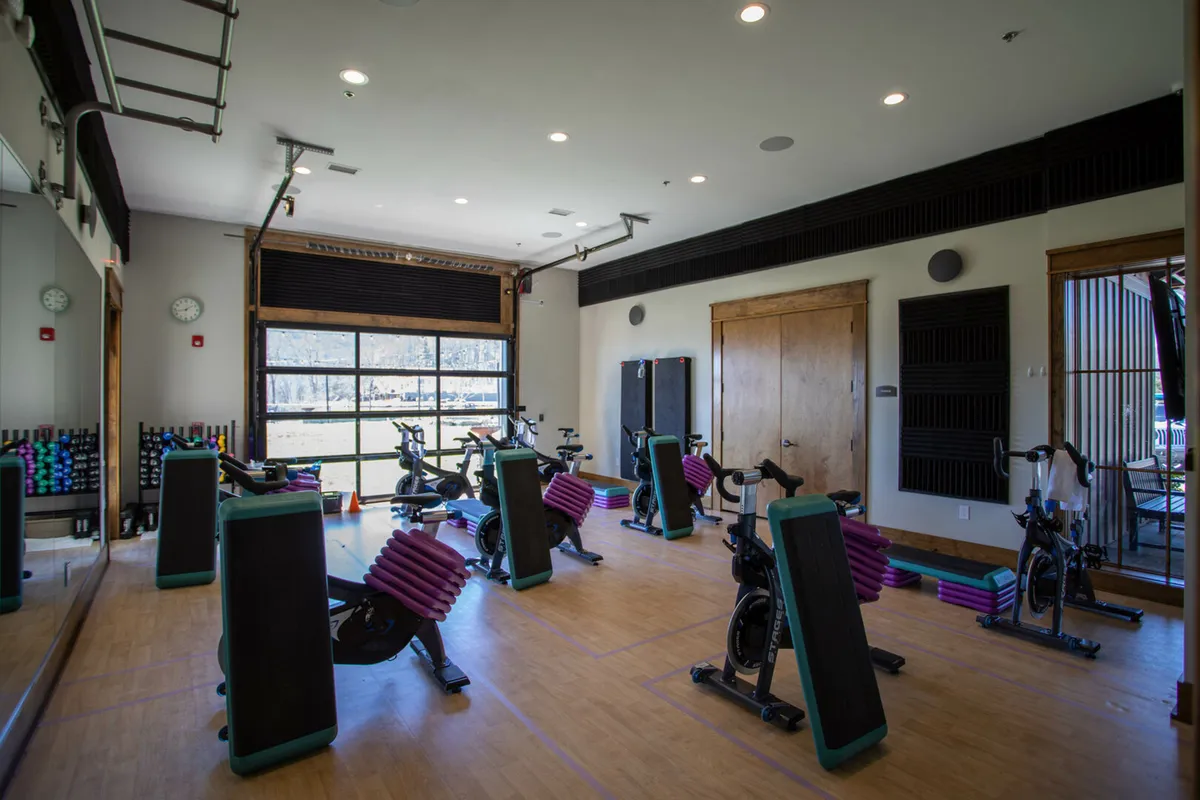 A bright, modern wellness center interior with rows of exercise bikes, a weights rack, and expansive windows letting in natural light. Groups participate in fitness classes and individuals pursue personal workouts, with an energetic, communal vibe.