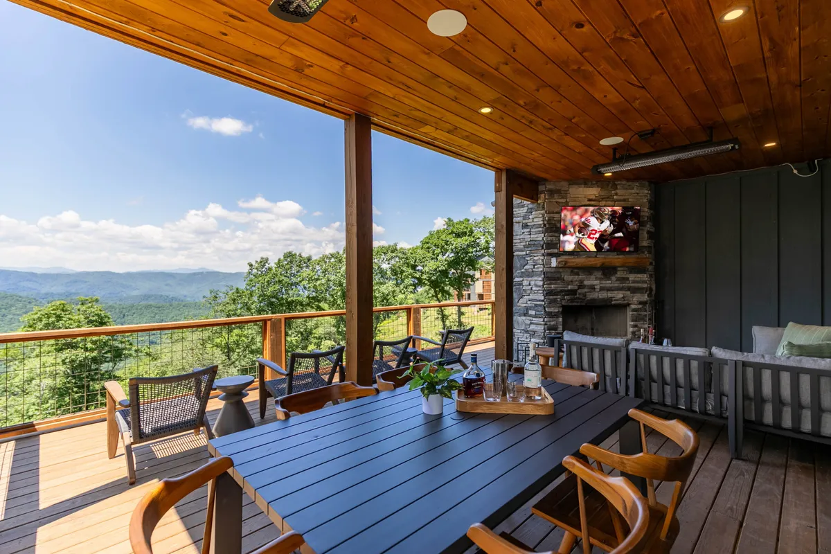 A spacious outdoor dining area with a large table, mountain views, a substantial outdoor fireplace, and a TV mounted on the wall. Friends or family can be imagined gathering here for meals or celebrations, with the mountains as a backdrop.