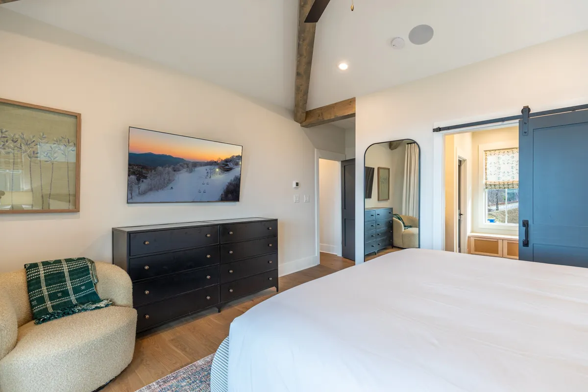 A large, comfortable bed is complemented by a TV displaying a scenic landscape, a dark dresser, and an accent chair. The room is styled in soothing colors, emphasizing both relaxation and modern amenities. Soft lighting and clean lines give the suite a tranquil, contemporary feel.