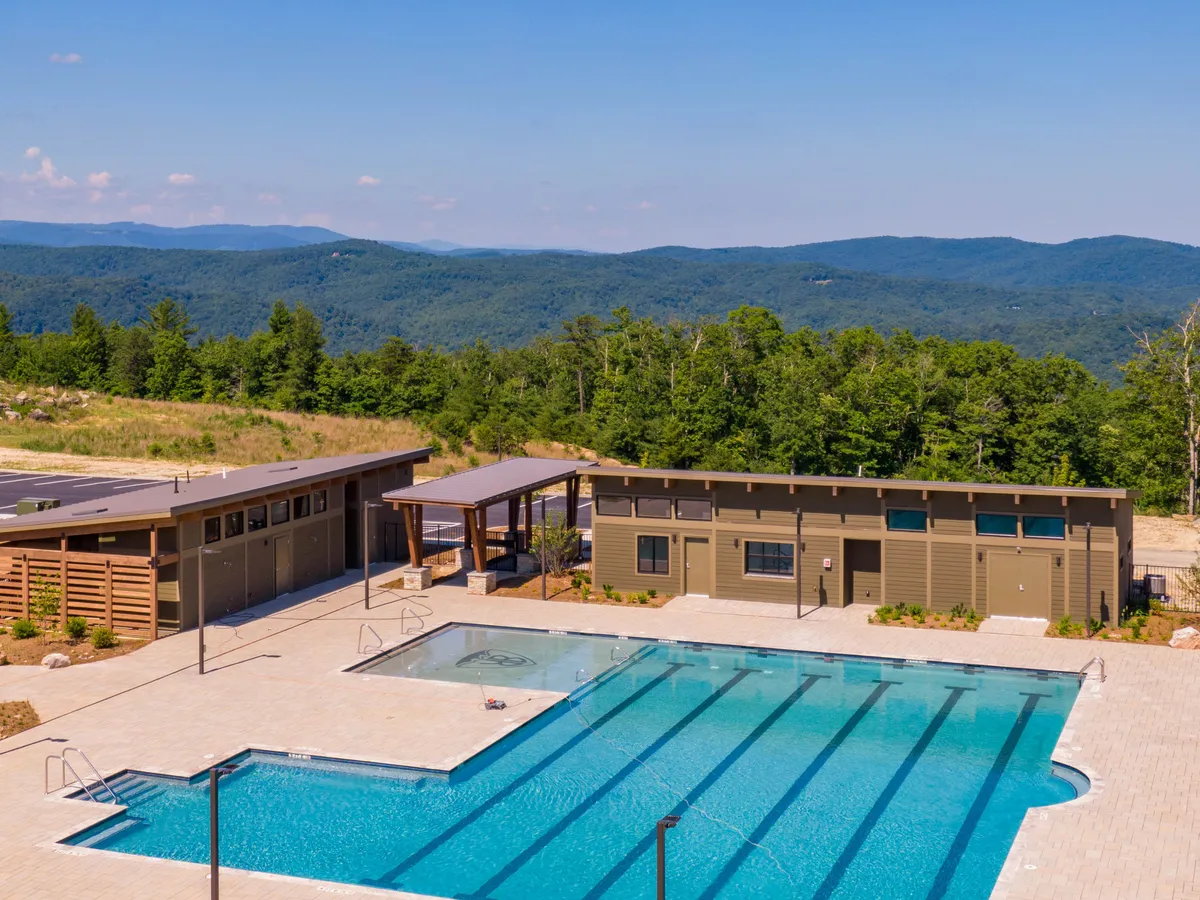 A vibrant outdoor swimming pool area surrounded by landscaped greenery and recreational buildings, with the majestic mountains in the background. The scene hints at opportunities for swimming, sunbathing, social gatherings, and outdoor events under open skies.