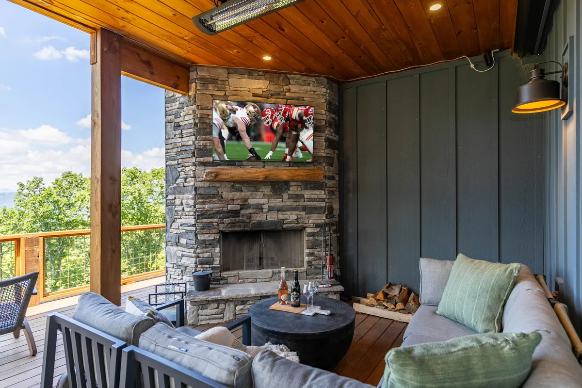 An inviting outdoor deck with a prominent stone fireplace, wall-mounted television, and comfortable seating. A wooden deck stretches out, offering both covered and open-air areas, perfect for gatherings and entertainment.