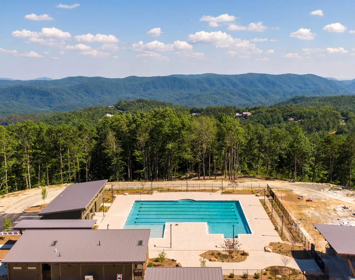 A spacious swimming pool is surrounded by lush greenery, modern amenity buildings, and scenic mountain backdrops. People are seen lounging, swimming, and socializing, highlighting the vibrant community aspect and luxury resort amenities available to residents and guests.