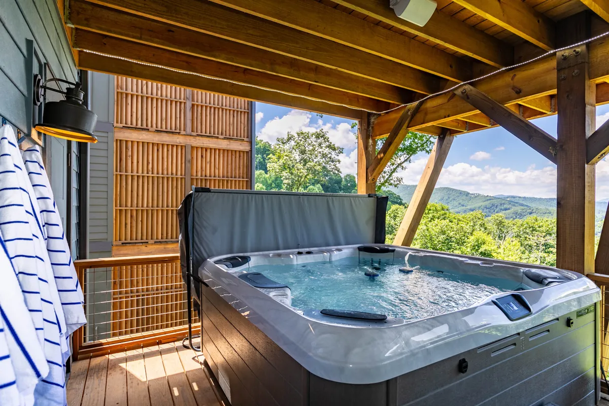 This image features a hot tub situated on a wooden deck framed by sturdy beams, with striped towels nearby and a sweeping mountain view in the background. The setting clearly conveys an outdoor relaxation space designed for enjoying both the view and the amenities.