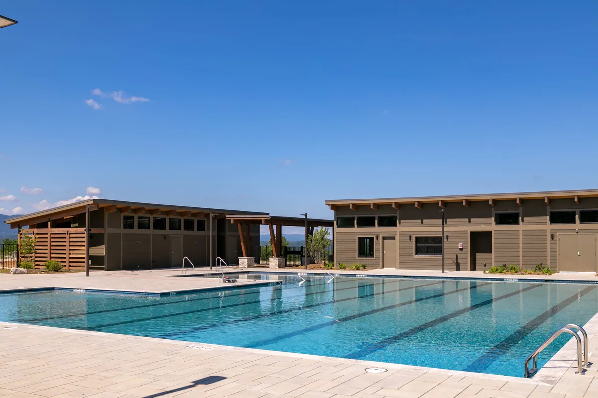 A stunning outdoor swimming pool surrounded by elegant resort buildings and lush landscaping. Residents lounge by the pool, swim, and gather for social activities, with rolling hills and sunset colors reflected in the water.