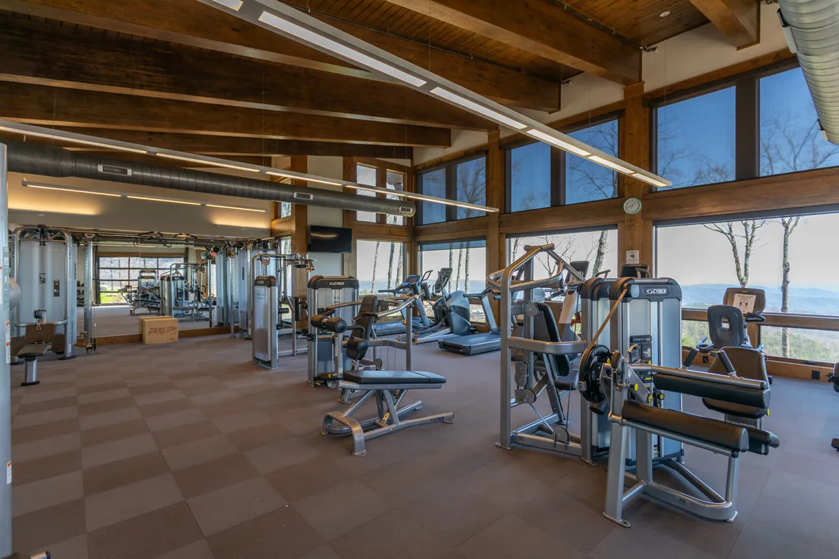 A bright, modern fitness center with exercise machines positioned near expansive windows that let in natural light and offer glimpses of the surrounding trees. Exposed wooden beams give the space a warm, welcoming feel, while the equipment showcases the Ascent Wellness & Fitness Center’s amenities.