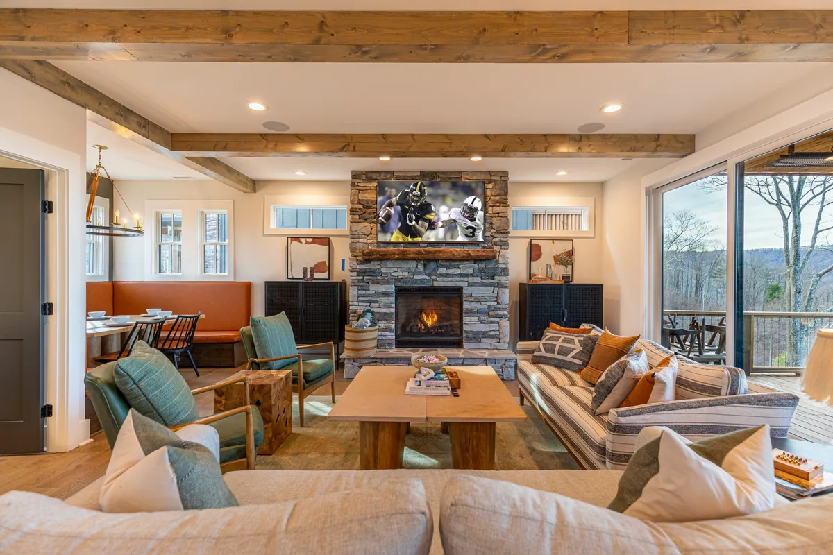 A spacious, vaulted living room with a striking stone fireplace as its focal point. Large windows frame panoramic mountain views, filling the space with natural light. A central coffee table is surrounded by plush seating, and a high-definition TV is mounted nearby, creating an inviting area for relaxation or entertainment.